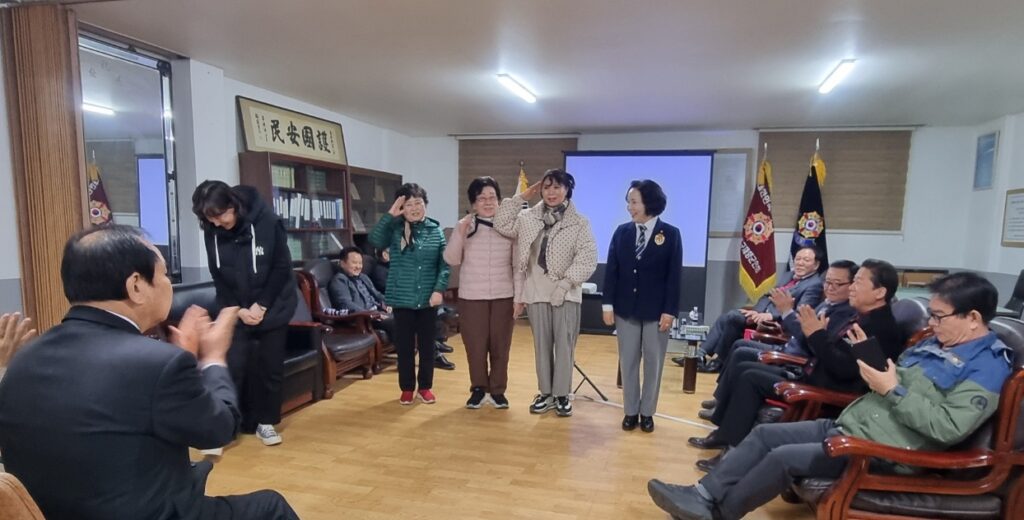 회의장 중앙에서 여성 회원들이 거수경례를 하며 인사를 나누고 이를 지켜보는 회원들이 박수를 보내는 장면