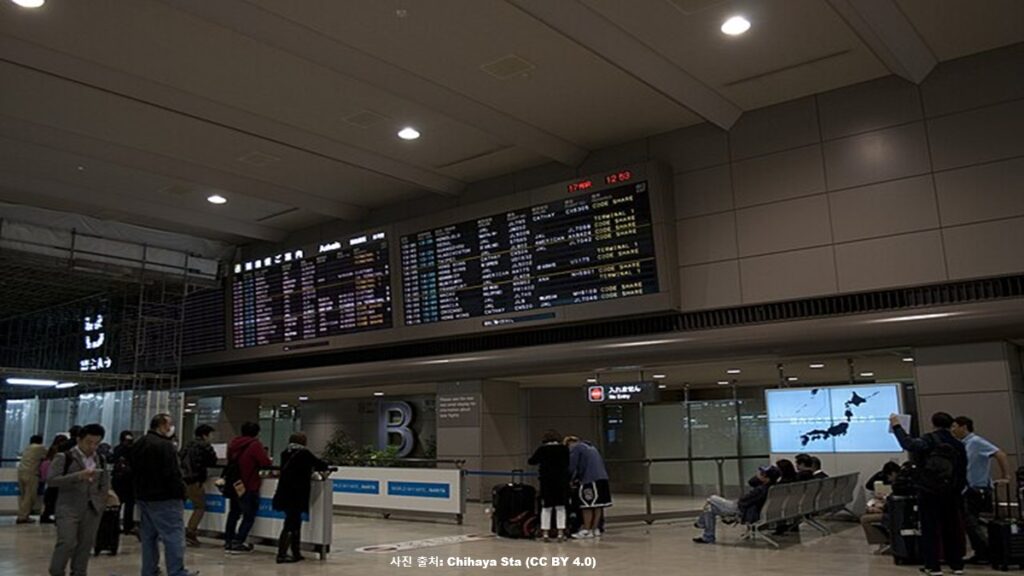 일본 나리타 공항 제1터미널 도착층의 어두운 실내에서 수많은 항공편 일정이 표시된 대형 전광판을 바라보며 이동하거나 대기 중인 여행객들과 가방을 챙기는 사람들의 모습. 미국의 비자 보증금 제도 확대 및 입국 심사 강화로 인한 여행객들의 불편과 공항의 긴장된 분위기 관련 국제 시사 보도 이미지. (출처: 위키미디어 공용 / 저작자 Chihaya Sta)