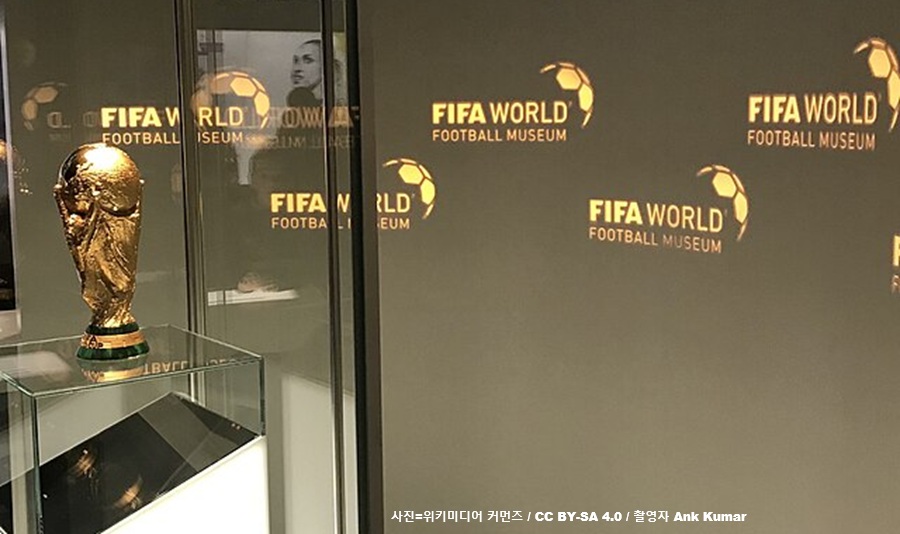 유리 전시관 안에 보관된 황금색 FIFA 월드컵 트로피와 그 뒤로 보이는 'FIFA WORLD FOOTBALL MUSEUM' 로고 벽면. 2026 북중미 월드컵의 기록적인 티켓 가격 폭등 및 FIFA의 상업주의 논란 관련 경제 보도 이미지. (출처: 위키미디어 커먼즈 / CC BY-SA 4.0 / 촬영자 Ank Kumar)